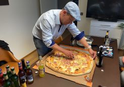 Pizzaanschnitt beim Treffen der Drupal User Group Bodensee am 17.6.2024 - Foto mit freundlicher Genehmigung von: Matthias Walti / mazze.ch Riesige Pizza auf der MItte eines Tisches, umgeben von Getränken. Die Pizza wird eben vom Gastgeber mit blauer Mütze geschnitten.