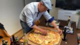 Riesige Pizza auf der MItte eines Tisches, umgeben von Getränken. Die Pizza wird eben vom Gastgeber mit blauer Mütze geschnitten. 